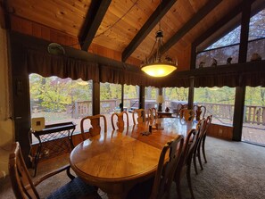 Zona pranzo in legno con un grande tavolo rotondo e sedie, un lampadario e vista su una terrazza con ringhiera.