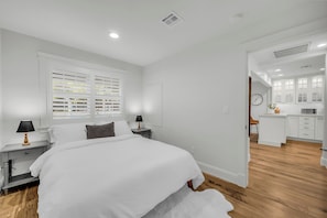 A modern bedroom with a large bed, bedside tables, a window with blinds, and a view into the kitchen.