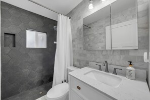 A modern bathroom with a hexagonal tile shower, a white toilet, a large mirror, and a white sink with a soap dispenser.
