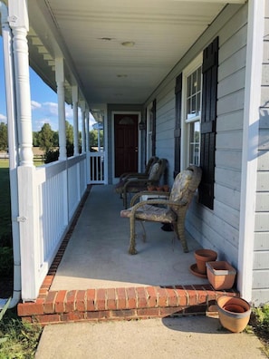 A porch with a brick step leading to a white door, two wicker chairs, and terracotta pots.