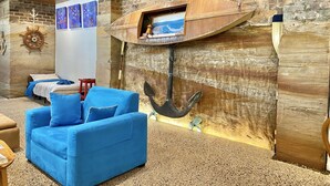 Habitación temática náutica, con un gran ancla y un timón de barco en la pared, un sofá azul y una pequeña mesa con una silla roja.