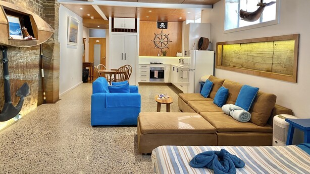 Una sala moderna con un sofá azul, un sofá marrón y una cama a rayas. Hay una cocina con estufa, refrigerador y un adorno en forma de timón de barco.