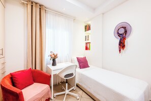 A small, well-lit room with a desk, chair, bed, and a bookshelf.