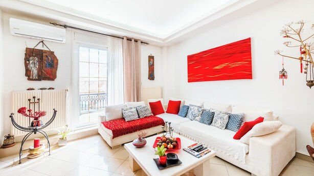 A modern living room with a white sofa, red cushions, a coffee table with decorative items, and a red abstract painting on the wall.