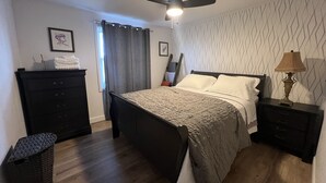 Une chambre à coucher comprenant un lit, une commode, une table de chevet, une lampe, un ventilateur de plafond et une fenêtre avec des rideaux.