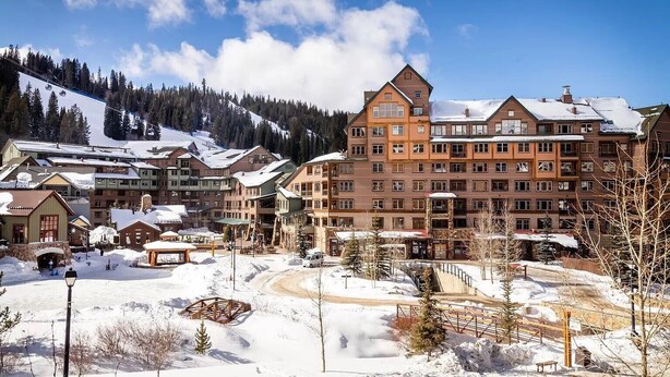 Un hotel de varios pisos con un telesilla de esquí al fondo, rodeado de árboles cubiertos de nieve y un cielo despejado.