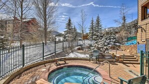 Un jacuzzi rodeado por un patio de ladrillo, con una cerca metálica y árboles nevados al fondo.