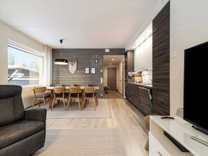 A modern living room with a sofa, a dining area with a table and chairs, a kitchen with a built-in oven, and a TV on a stand.