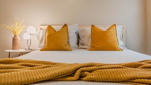 Una cama bien hecha con una manta amarilla, dos almohadas y una mesita de noche con un florero de plantas secas y una lámpara.