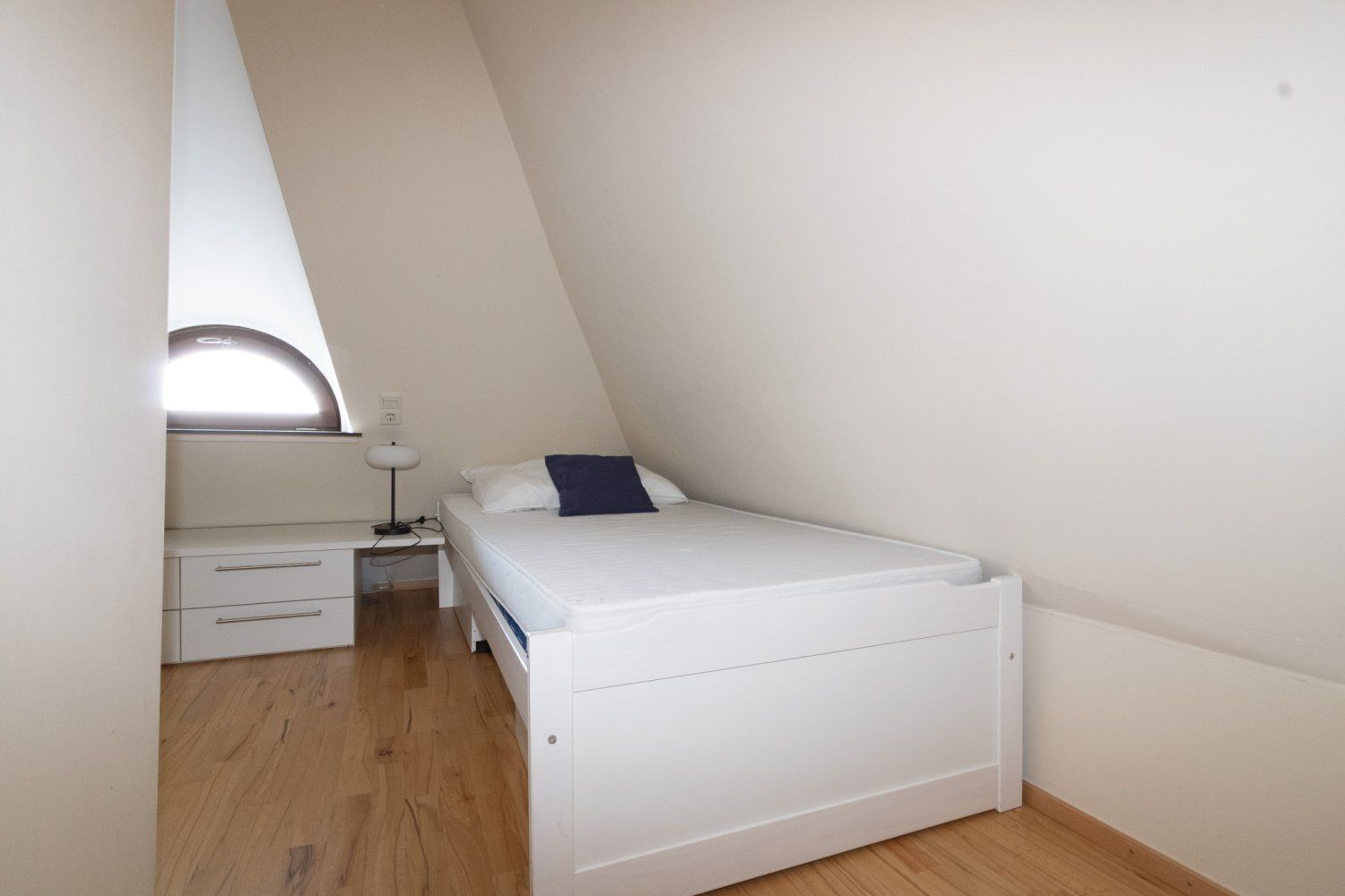 A small, white, single-bed room with a desk and a lamp.