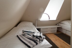 Ein kleines, modernes Schlafzimmer mit schrägem Dach, einem Bett mit gestreifter Decke, einem Nachttisch mit Schubladen und einem Fenster, das für natürliches Licht sorgt.