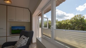 A room with a TV on a cabinet, a chair with a patterned cushion, and a large window with blinds. - Reetdachhaus Süderende (Hörnum (Sylt))