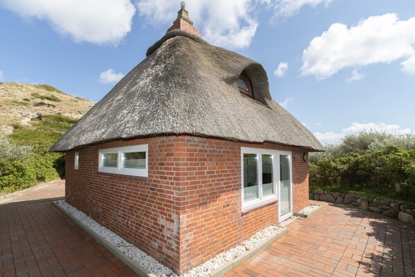 A thatched-roof cottage with a brick exterior, white-framed windows, and a stone foundation.