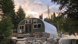 A rustic cabin with a curved roof, a wooden deck, and a stone foundation. - Luxurious Quonset hut in the woods, mountain views (Kila)