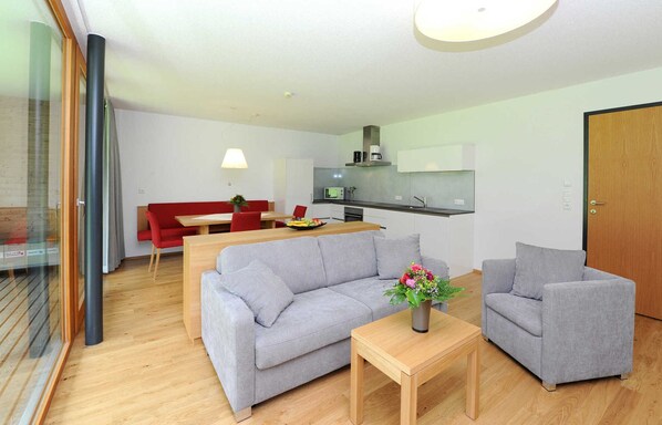 A modern living room with a grey sofa, a wooden coffee table, and a red dining table. A kitchen with a microwave and stove is visible in the background.