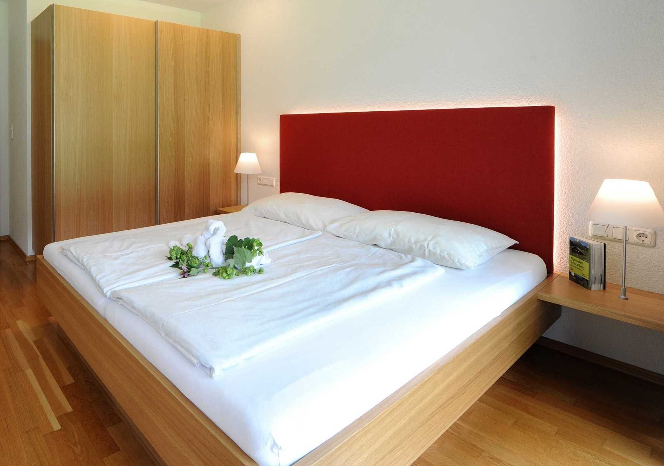 A neatly made bed with white linens, a red headboard, and a bedside table with a lamp.