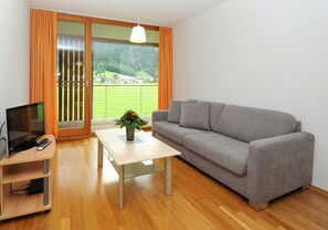 Ein modernes Wohnzimmer mit einem grauen Sofa, einem Holztisch, einem Fernseher auf einem Lowboard und Blick auf eine grüne Landschaft durch große Fenster.