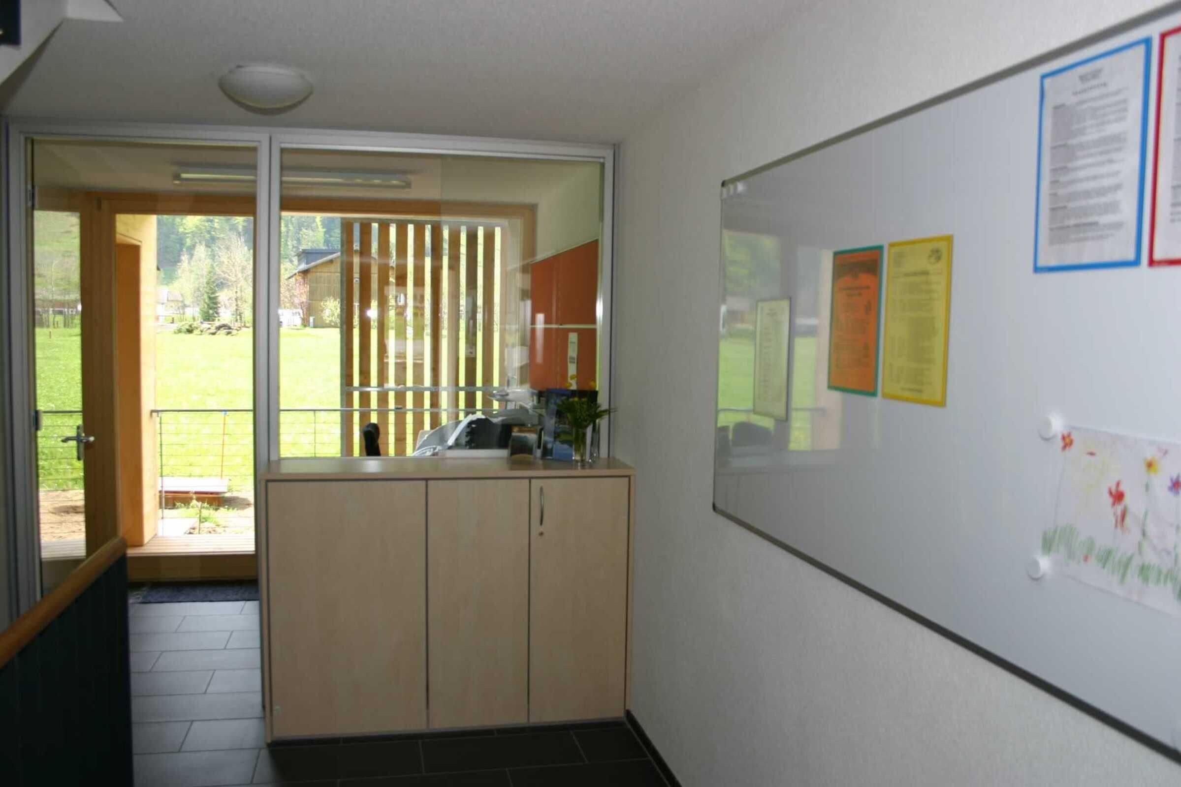 A room with a sliding glass door leading to a balcony, a whiteboard with notices, and a wooden cabinet.