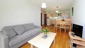 A living room with a grey sofa, a wooden dining table with chairs, a TV on a stand, and a kitchen in the background. - Apartment Diedams [ground floor | 45 m²] - Apartments Susanne (Rehmen)