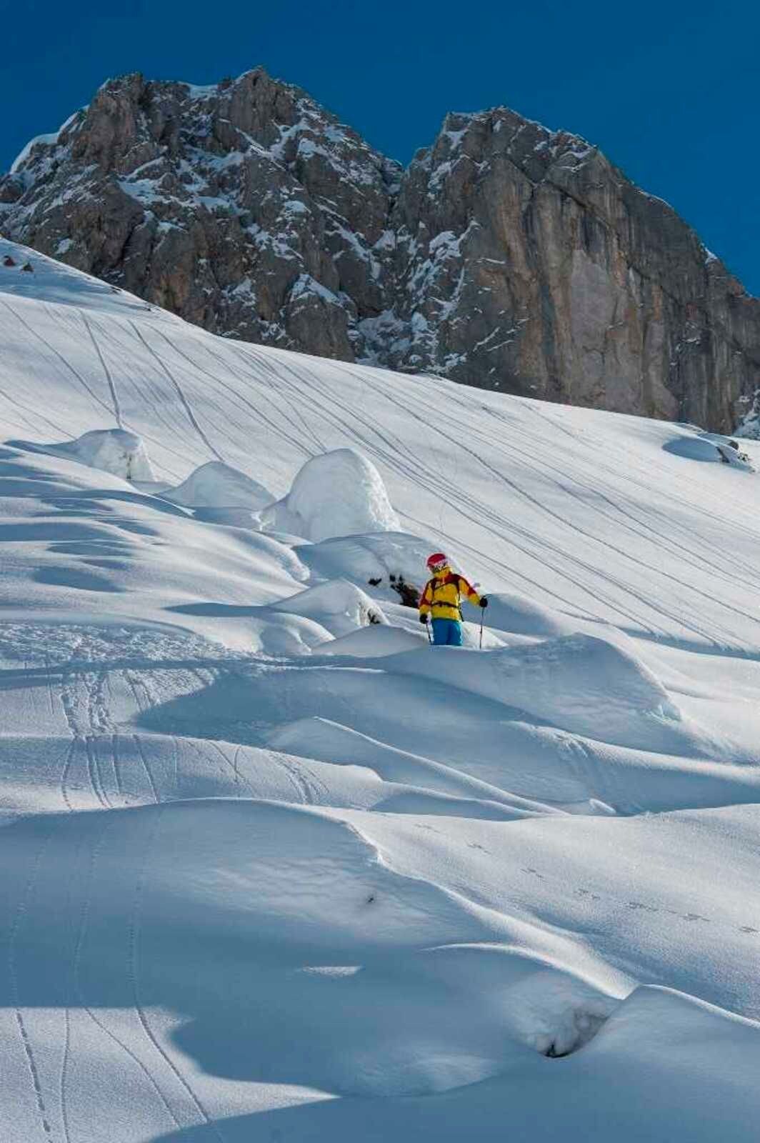 Uma pessoa esquiando em uma encosta coberta de neve, com montanhas rochosas ao fundo.