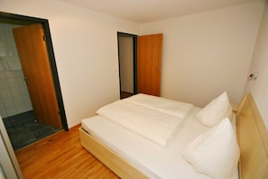 A bedroom with a bed, wooden flooring, and a door leading to another room.