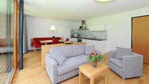 Una sala de estar moderna con un sofá gris, una mesa de centro de madera y una mesa de comedor roja. Se ve una cocina con microondas y estufa al fondo.
