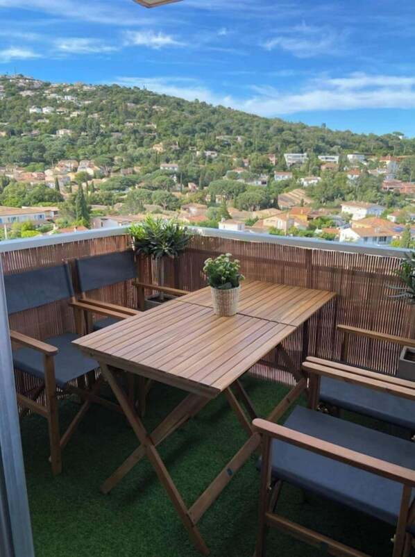 Une terrasse en bois avec une table et des chaises, entourée d’une clôture en bambou, donnant sur une colline où se trouvent des maisons.