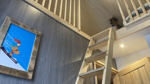 Eine Holztreppe mit Handlauf, ein gerahmtes Bild an der Wand und ein Erkerbereich mit einer Leiter.