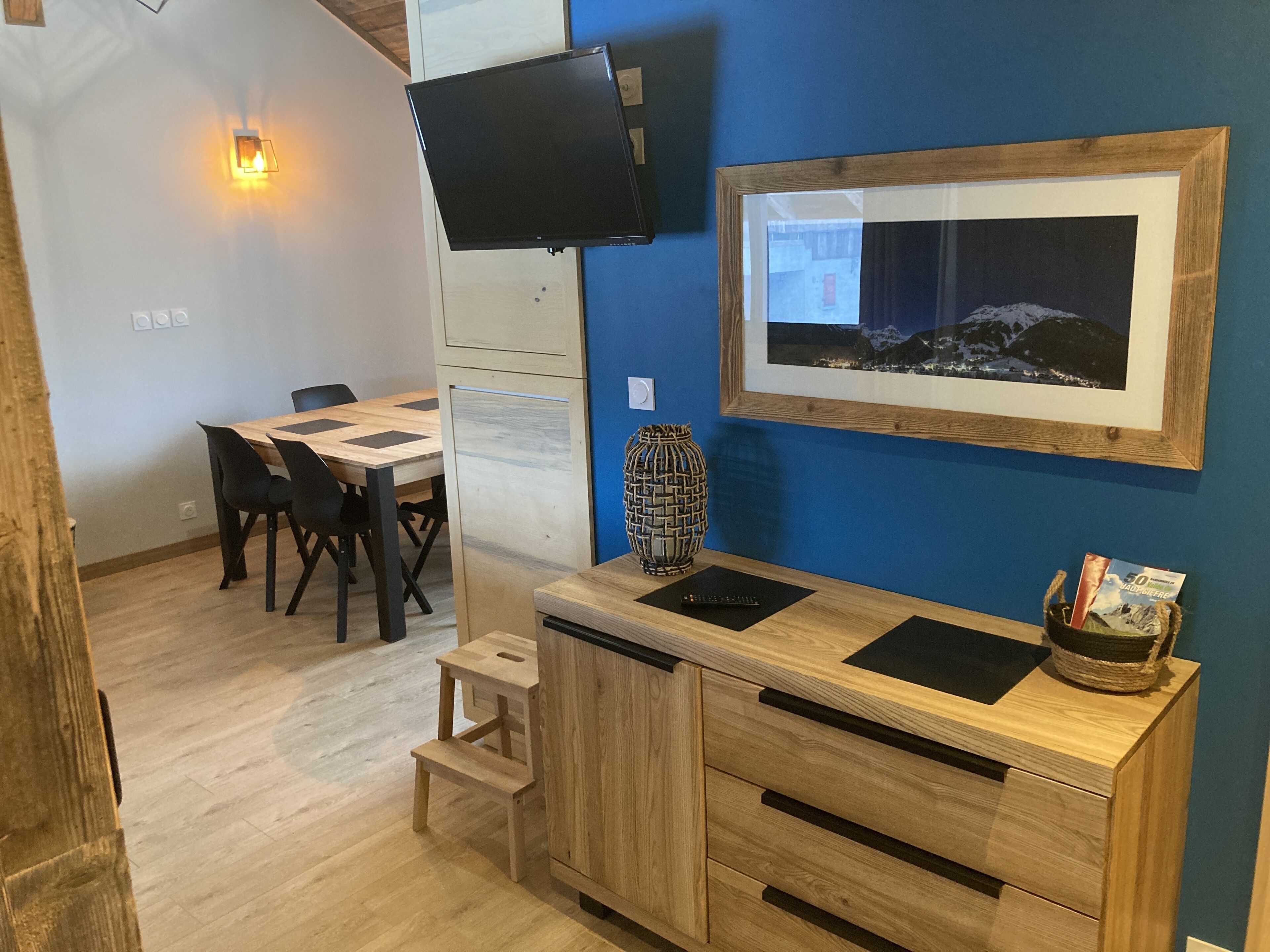 A modern room with a wooden desk, a flat-screen TV mounted on the wall, a framed picture, and a small wooden stool.