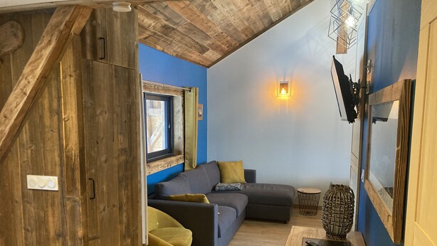 Ein gemütliches Loft-Zimmer mit Holzbalkendecke, einem grauen Sofa, einem kleinen Tisch und einem Fenster mit Vorhängen.