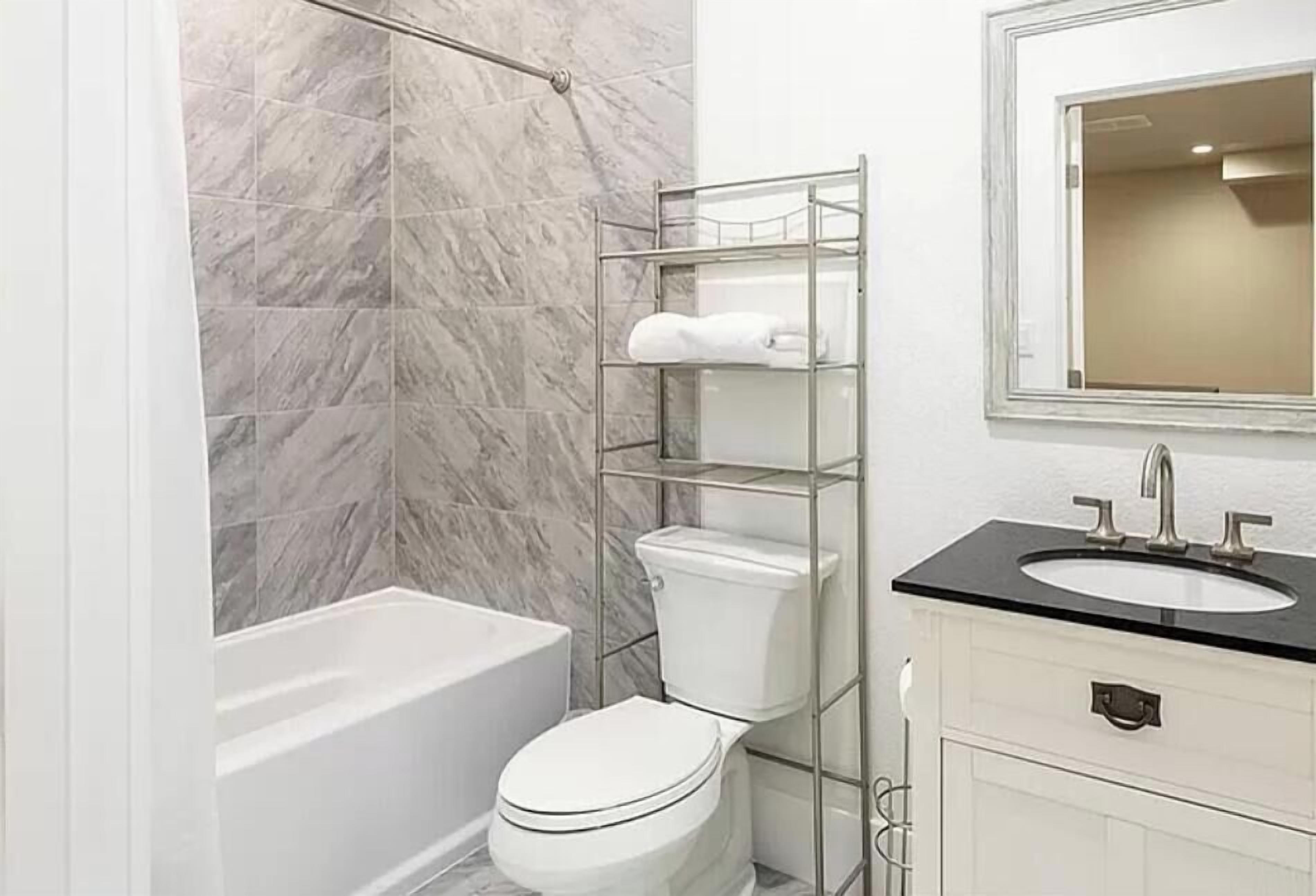Ein Badezimmer mit Marmorwand, einer Badewanne, einer Toilette, einem Metallregal mit Toilettenartikeln, einem Waschbecken und einem Spiegel.