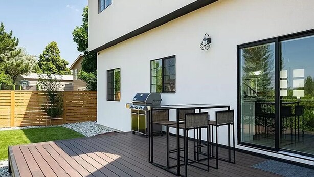 A modern outdoor patio with a wooden deck, a barbecue grill, and a dining table with chairs.