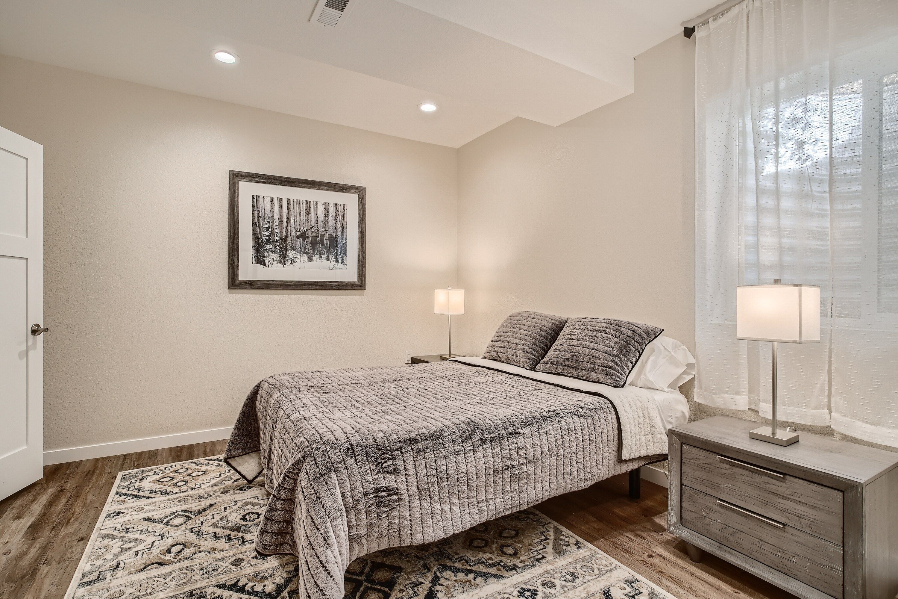 Un dormitorio con cama, lámparas de noche, mesita de noche, alfombra y un cuadro en la pared.