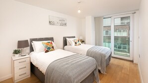 A modern bedroom with two beds, a nightstand, a lamp, and a window with a view of buildings. - Skyvillion Apartments - Barnet - Beautiful 2Bed Apartment with Balcony (New Barnet)