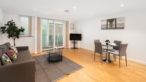 A modern living room with a sofa, a dining table with chairs, a TV, and a large window with a view of the outside. - Skyvillion Apartments - Barnet - Beautiful 2Bed Apartment with Balcony (New Barnet)