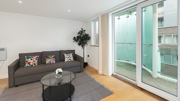 A modern living room with a grey sofa, colorful cushions, a glass coffee table, and a large sliding glass door leading to a balcony. - Skyvillion Apartments - Barnet - Beautiful 2Bed Apartment with Balcony (New Barnet)