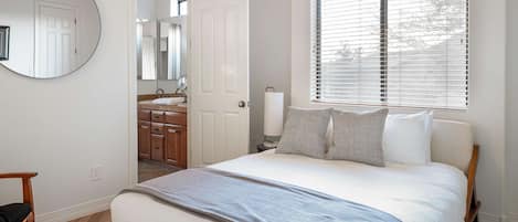 A bedroom with a bed, a bedside table, a mirror, a window with blinds, and a view of the bathroom.
