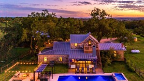 A large house with a stone exterior, a swimming pool, and a patio area with outdoor seating. - Grand Oaks Ranch · Pool·Spa · Game Barn · Animals (Fredericksburg)