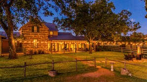 A stone building with a porch and string lights, surrounded by a fenced yard with trees. - Grand Oaks Ranch · Pool·Spa · Game Barn · Animals (Fredericksburg)