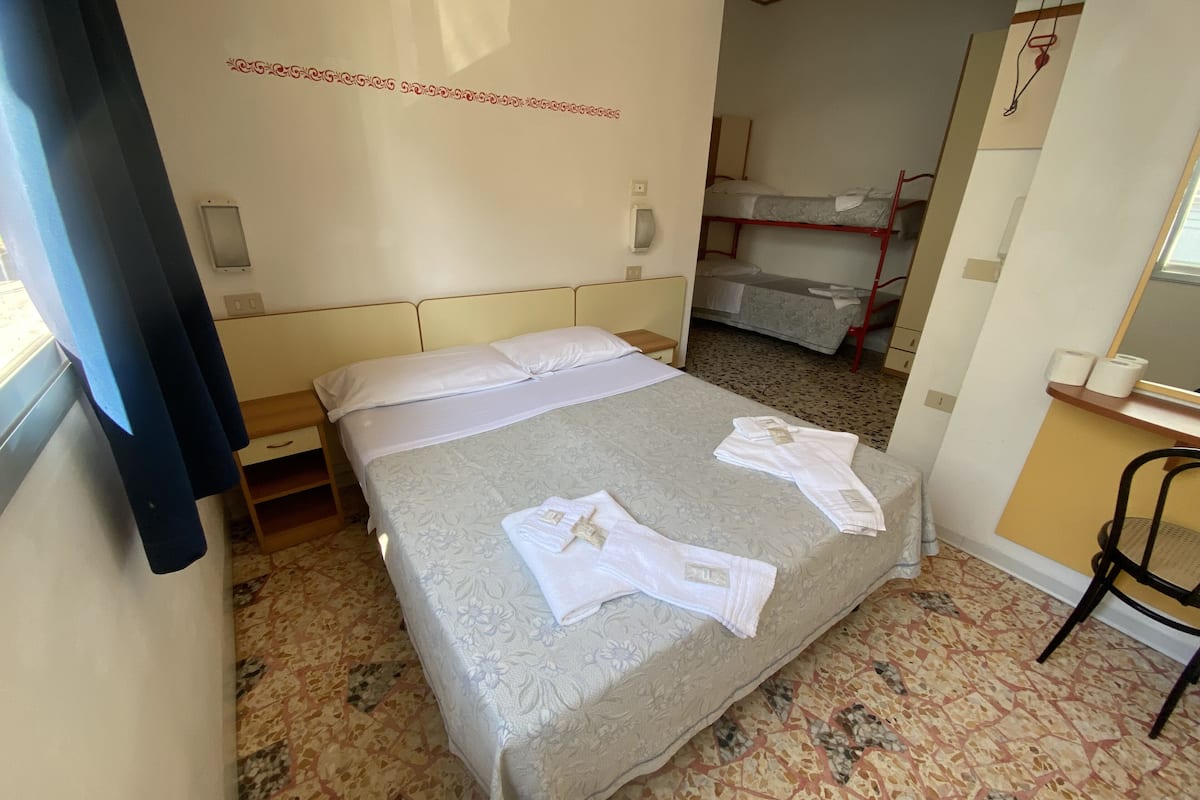A hotel room with a bed, a desk, a chair, and a bunk bed in the background.