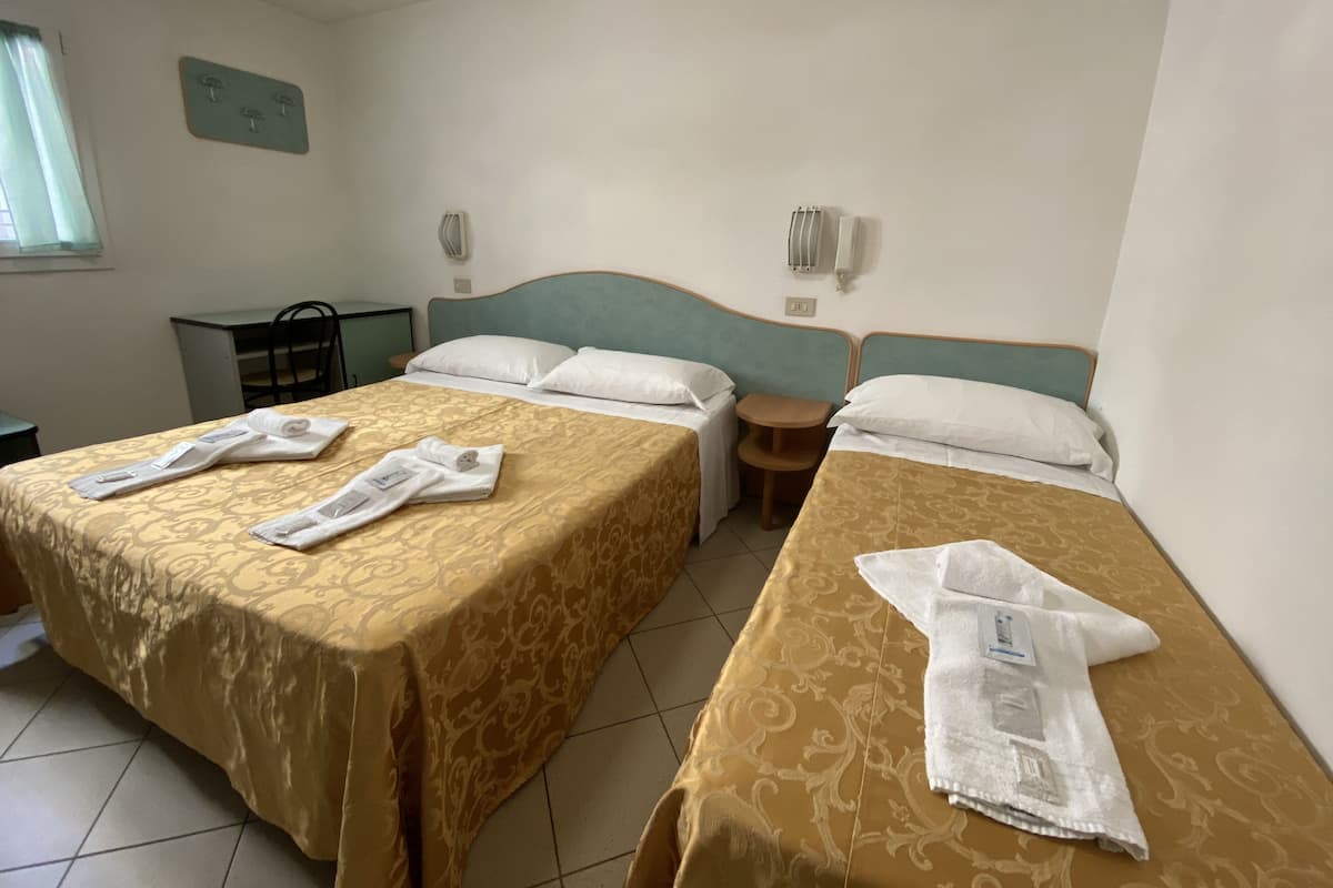 A hotel room with two beds, each with a patterned bedspread and white pillows. There is a small wooden table between the beds, and a desk with a chair to the left.