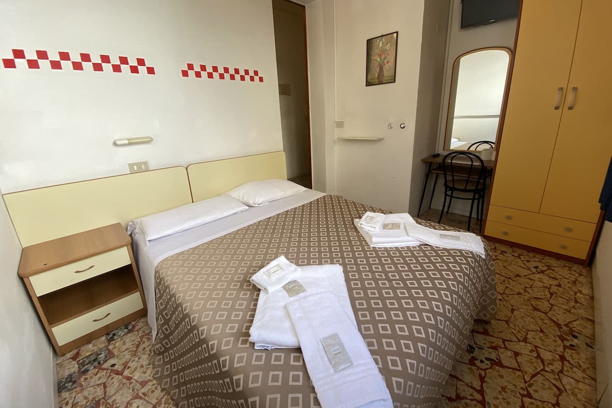 A neatly made bed with a patterned bedspread, a bedside table with a drawer, a small shelf, and a mirror on the wall.