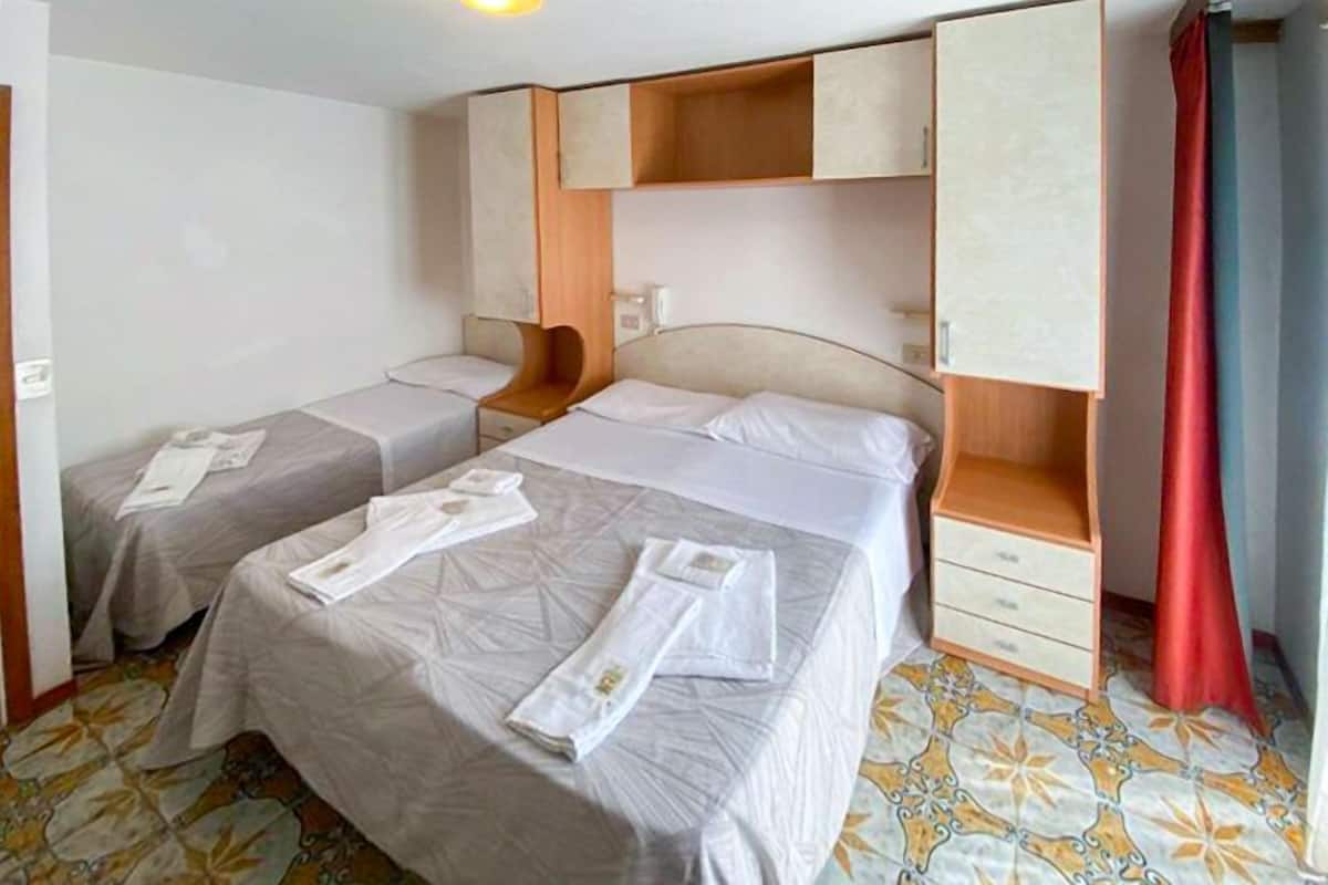 A hotel room with two beds, a wooden wardrobe, a red curtain, and patterned floor tiles.