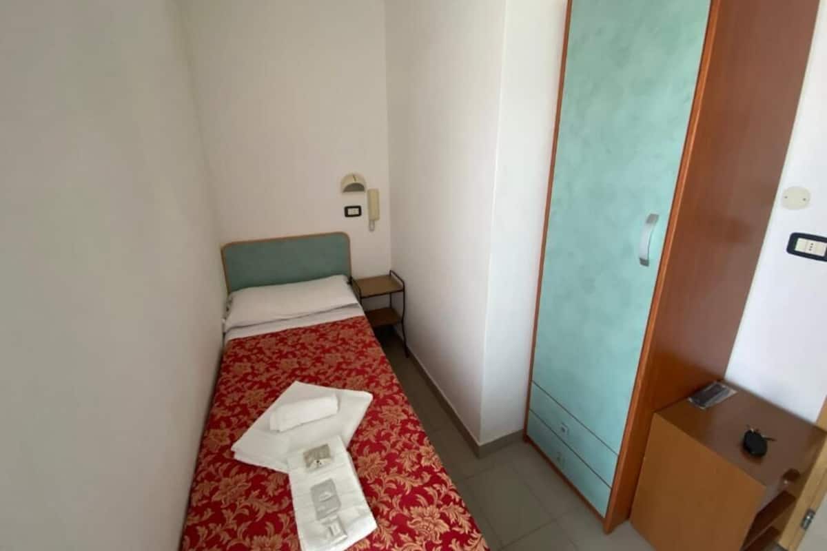 A small, simple room with a bed, a wardrobe, a bedside table, and a wall-mounted telephone.