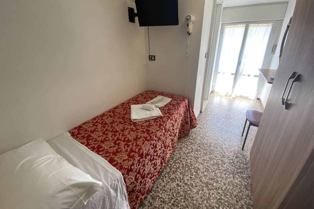 A small, single-bed room with a red floral bedspread, a white pillow, a TV mounted on the wall, and a door leading to a balcony.