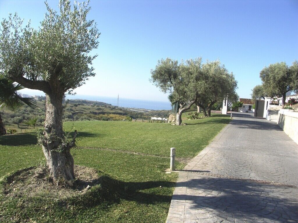 A paved pathway leading through a grassy area with trees, overlooking a distant landscape.