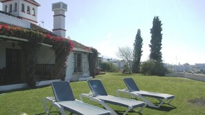 A building with a white facade and red-tiled roof, a chimney, and a balcony with red flowers. There are three white lounge chairs on a green lawn. - Spectacular villa with private pool and beautiful views (Nerja)