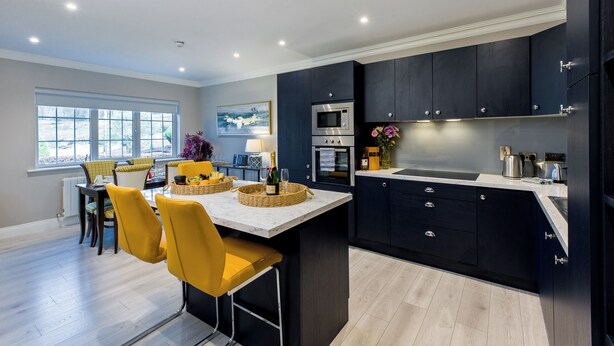 Une cuisine moderne avec des meubles de couleur sombre, un îlot central et un coin repas avec des chaises jaunes.