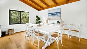 A dining area with a white table and chairs, a large window with a view of greenery, and a painting of the ocean on the wall. - Sandy Feet Retreat in Cape Woolamai (Cape Woolamai)