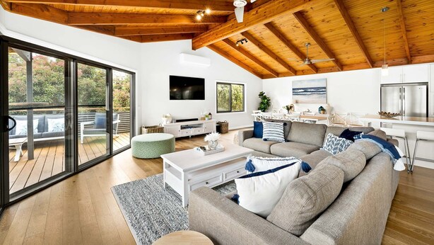 A spacious living area with a large sectional sofa, a coffee table, and a television. There is a sliding glass door leading to an outdoor deck. - Sandy Feet Retreat in Cape Woolamai (Cape Woolamai)